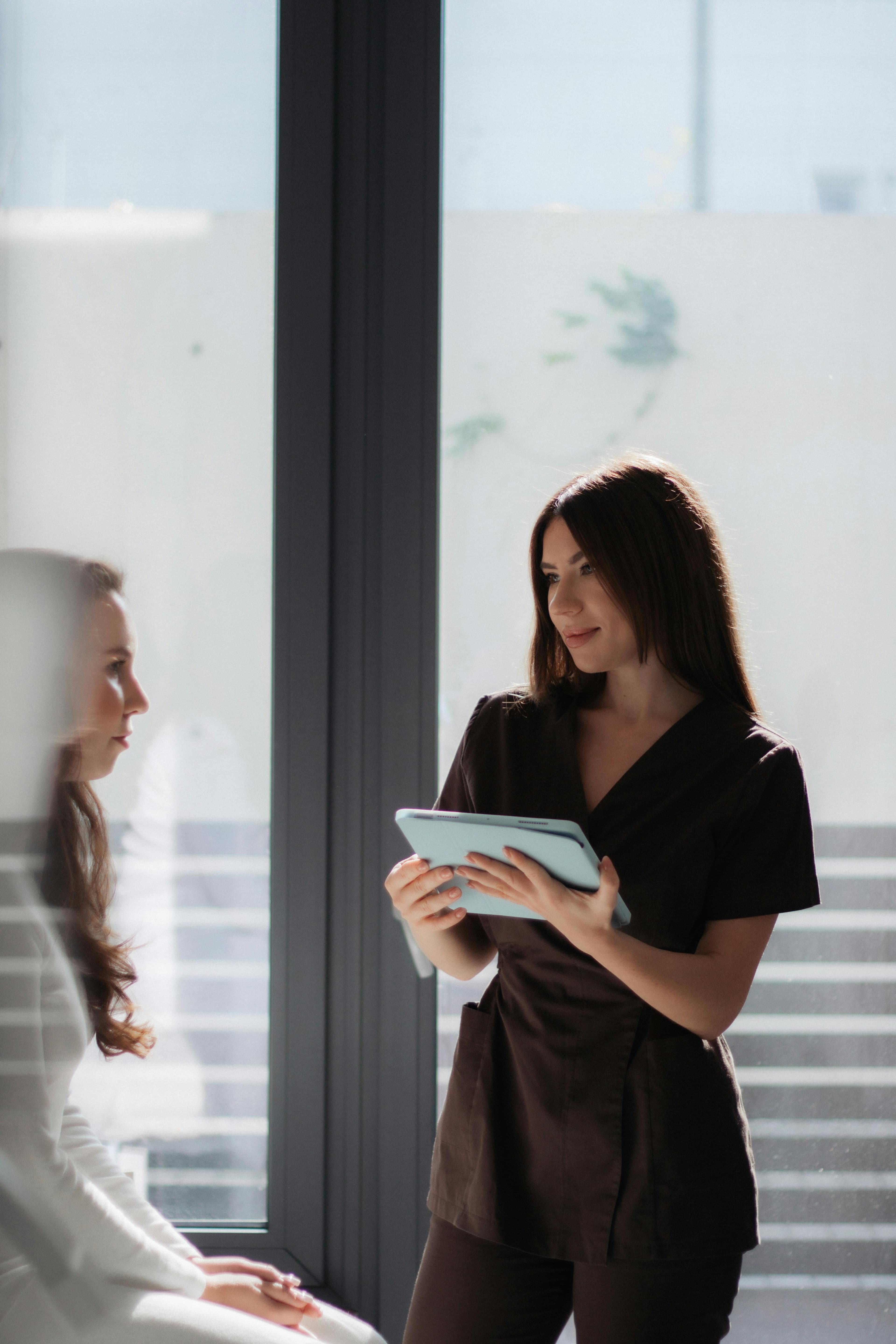 A professional woman in brown scrubs holding a blue tablet while consulting with a client in a bright, modern office setting.